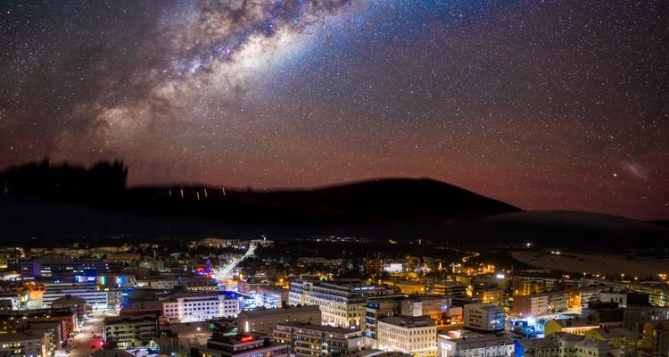 Ciel nocturne parsemé d'étoiles et Voie lactée s'arquant au-dessus d'un paysage urbain brillamment illuminé.