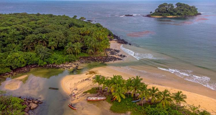 Vista aérea de una playa tropical con palmeras, laguna y pequeños botes de pesca, islote en el horizonte.