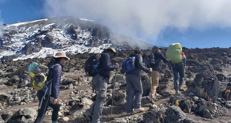 Une file de randonneurs gravit une pente volcanique rocheuse en direction d'un sommet enneigé au milieu de nuages flottants.