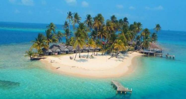 A tiny palm-fringed island surrounded by turquoise waters and a wooden pier extending over a coral lagoon.