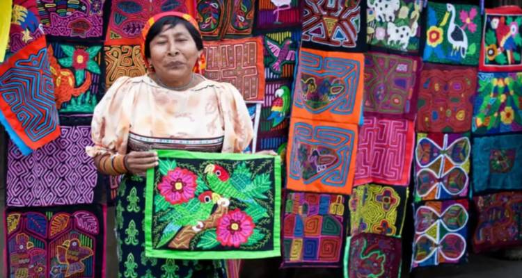 A Guna artisan proudly displays a colourful mola textile against a vibrant backdrop of embroidered fabrics.