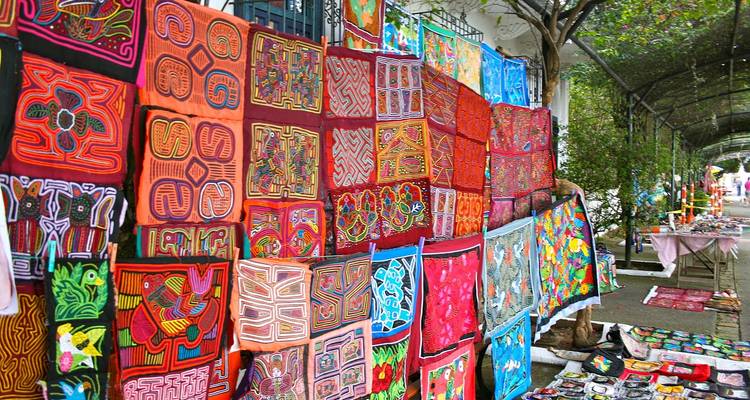 A colorful outdoor display of traditional hand-stitched textiles and crafts lining a shaded street market.