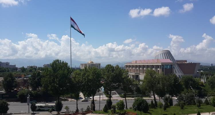 Large national flag of Tajikistan flies high over a parkland vista and modern civic buildings.