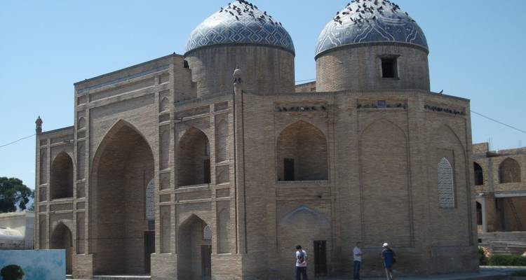 Brick mausoleum crowned by twin blue-tiled domes dotted with pigeons under a bright sky.