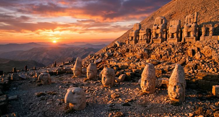 Ancient stone heads and seated statues of Mount Nemrut glowing orange at sunset over distant mountains.