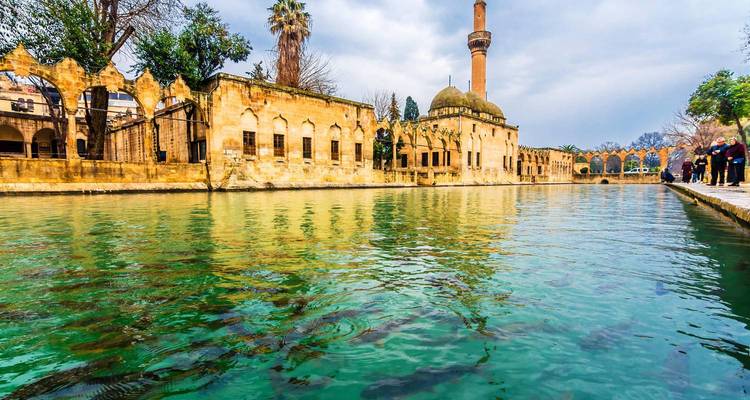 Golden-hued mosque and colonnades reflected in the green waters of Balıklıgöl with visitors along the bank.