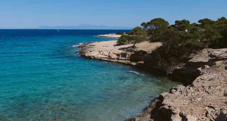 L'eau turquoise cristalline vient lécher le rivage rocheux calcaire bordé de pins et d'îles lointaines.