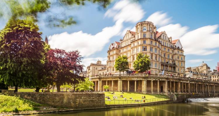 Le grand manoir en bordure de rivière et ses terres paysagées luxuriantes à Bath baignent sous un ciel dégagé.