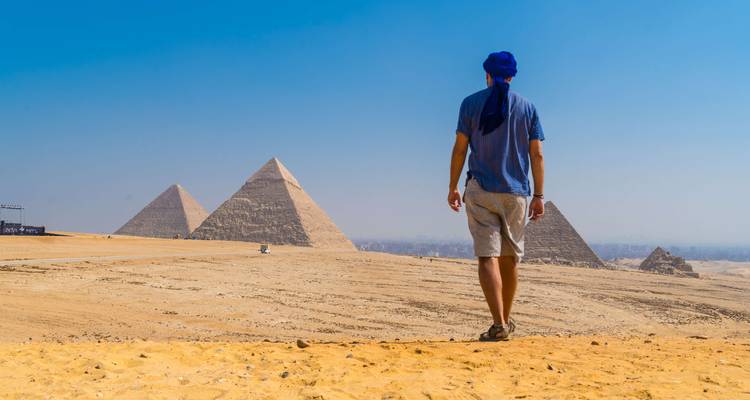 Lone traveler walking across the desert toward the iconic Pyramids of Giza under a clear blue sky.