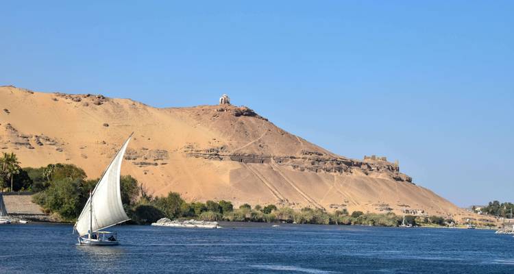 Traditional felucca sailboat gliding along the Nile with sandy hills and ruins in the background.