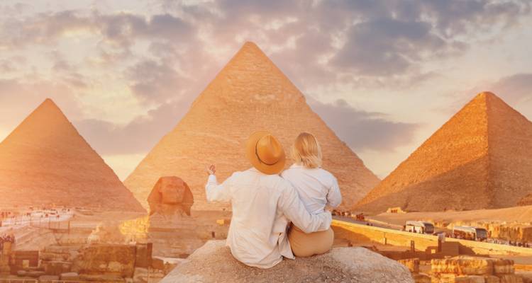 A couple sits on a rock admiring the Pyramids and Sphinx bathed in warm sunset light.