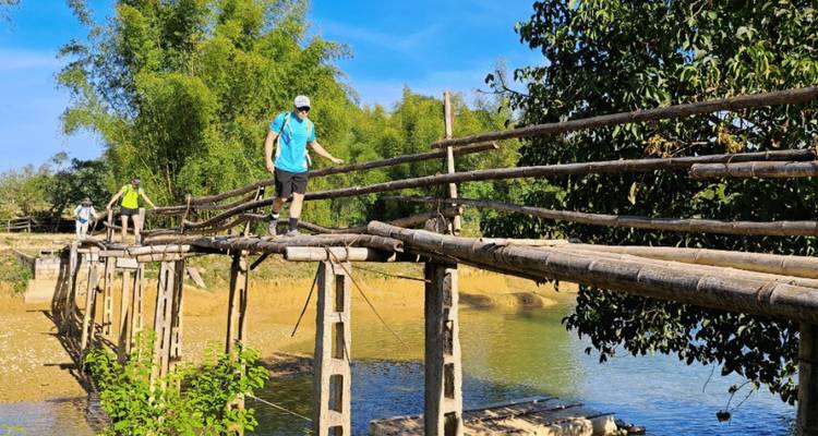Un pequeño grupo equilibrándose cuidadosamente sobre un puente de madera rústico que cruza un río poco profundo bajo un cielo azul.