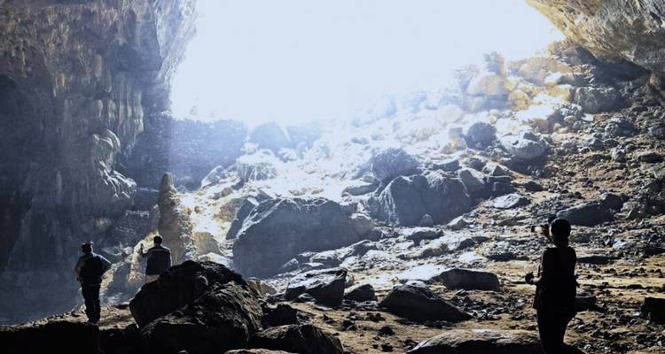 Exploradores recortados contra la luz dentro de una enorme cámara de cueva con luz natural brillante entrando por la entrada.