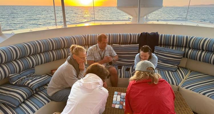 Group of travellers playing cards on cushioned deck seating as the sun sets over the water.