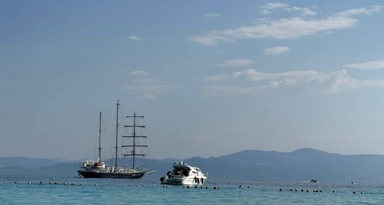 Two anchored yachts and distant mountains seen over calm blue water under light clouds.