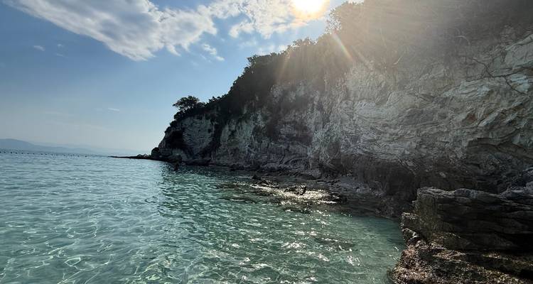 Sunlight breaking over a rocky cliff and crystal-clear turquoise water near a quiet cove.