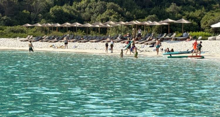 Pebble beach with rows of sunbeds, umbrellas and holidaymakers enjoying the shallow emerald water.