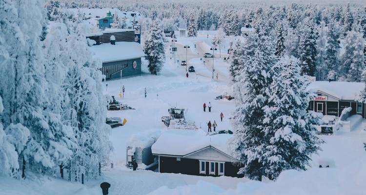 Village et forêt enneigés en Laponie finlandaise avec des personnes marchant parmi les congères profondes et les cabanes.