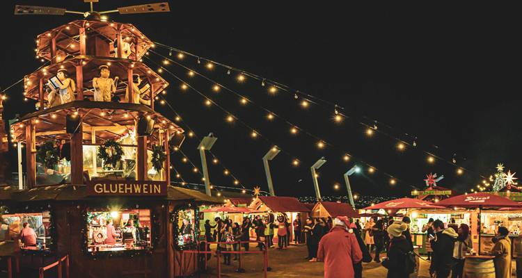 A festive Christmas market at night with twinkling lights, a tall wooden pyramid stall and visitors buying glühwein.