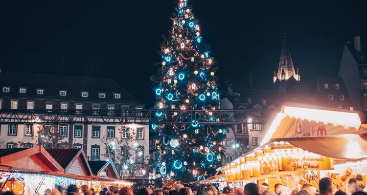 A towering illuminated Christmas tree dominates a busy night market square filled with wooden stalls and people.