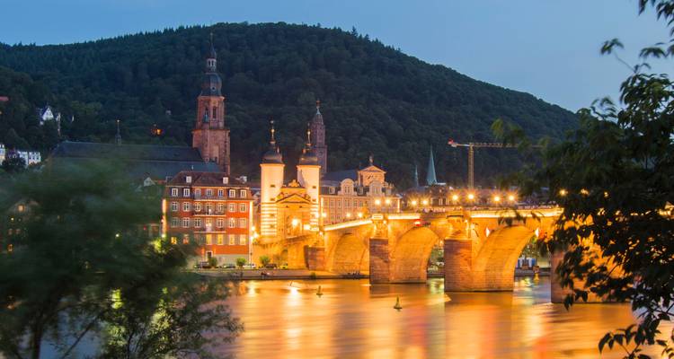 Illuminated stone arch bridge and church reflected on calm river at dusk in Heidelberg with forested hill backdrop