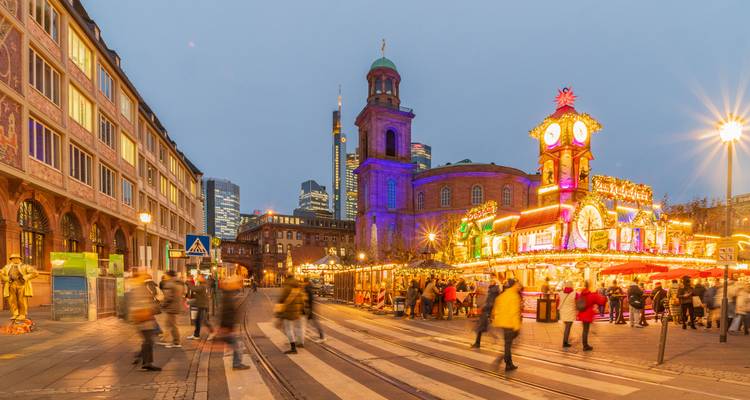 Evening scene of Frankfurt Christmas market with brightly lit stalls, St. Paul’s Church and modern skyline in background