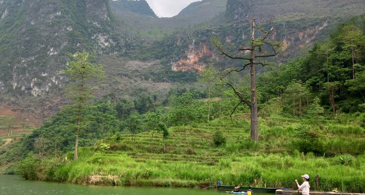 Un navegante local maneja una pequeña embarcación cruzando un río verde y tranquilo rodeado de colinas en terrazas y bosque.