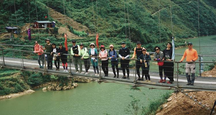 Un gran grupo de excursionistas posa en un estrecho puente colgante sobre un río de color jade en un exuberante valle.