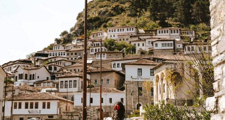 Traditional white Ottoman houses of Berat climb a hillside while a lone visitor walks below.