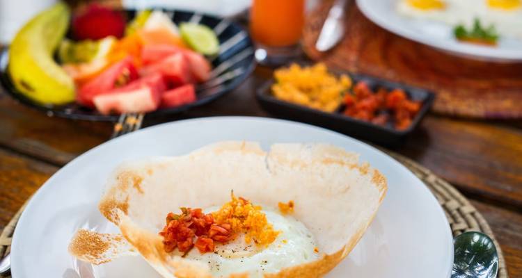 Sri Lankan breakfast of egg hopper topped with sambol on a white plate, with fresh fruit in background.
