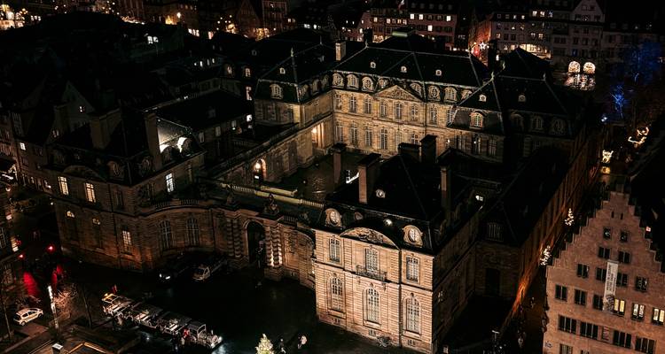 Vue aérienne nocturne d'un complexe palatial baroque orné illuminé par des lumières chaudes parmi les toits de la ville.