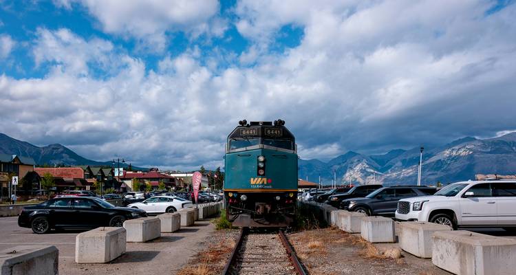 Blue VIA Rail locomotive on tracks amid parking lot with dramatic mountain backdrop.