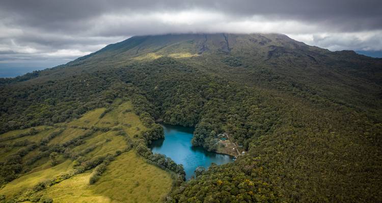 Aerial shot of a turquoise crater lake hidden within lush slopes of a cloud-capped volcano.