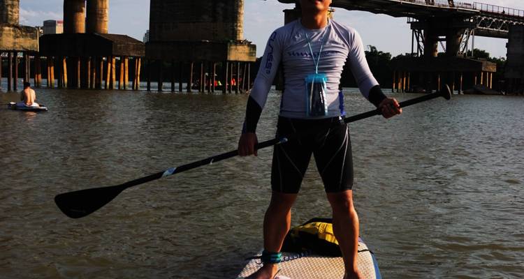Close-up van een man die balancerend op een paddleboard staat, met brugpijlers achter hem.