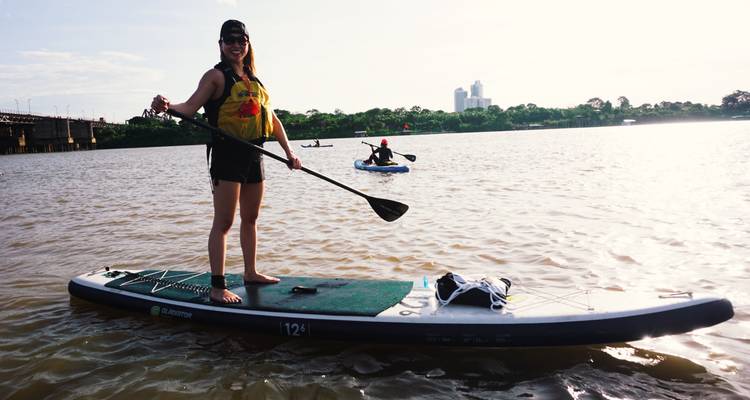 Vrouw die peddelt op kalm rivierwater met stadsskyline in fel tegenlicht.