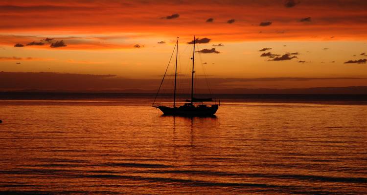 Silhouette d'un voilier solitaire ancré sur une eau calme face à un coucher de soleil d'un orange éclatant.
