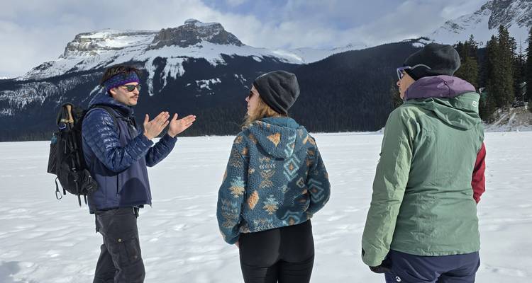 A guide talks to two travellers standing on a frozen lake with snowy mountains in the background.