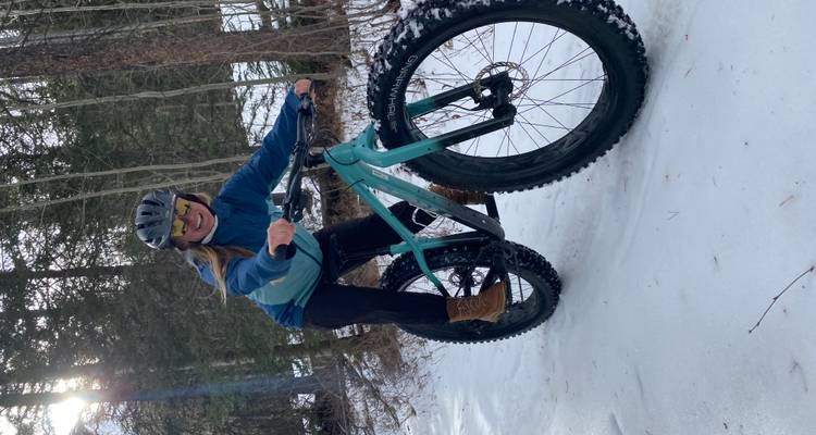 A woman rides a fat-tire bike on a snowy forest trail, smiling at the camera.