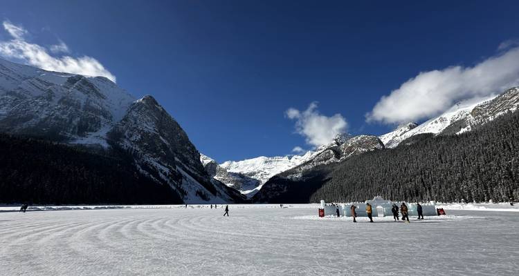 People skate on a vast frozen lake surrounded by dramatic snow-covered peaks and blue sky.