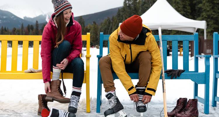 Two friends sit on colorful benches lacing up skates beside a snowy outdoor rink.