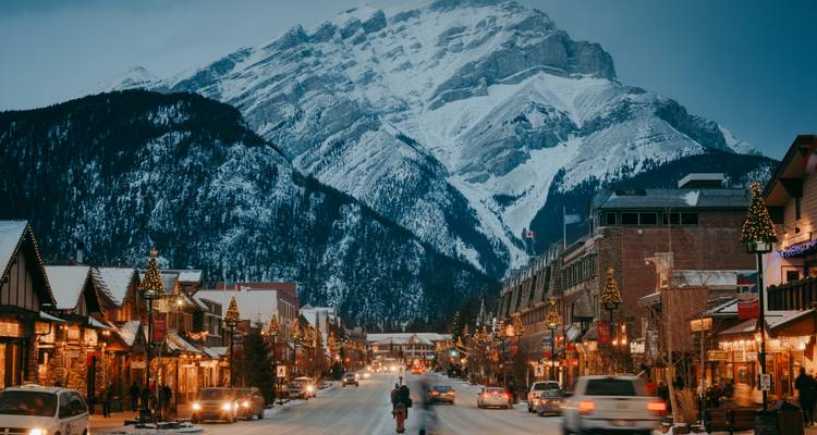 Evening lights glow along a charming alpine main street beneath a towering snowy mountain backdrop.