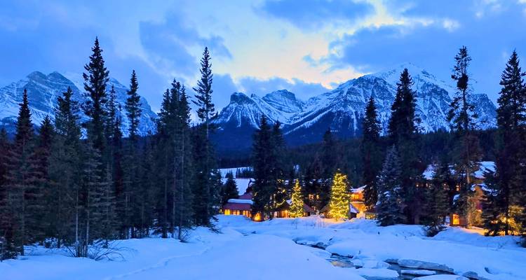 Cozy cabins and glowing trees sit in a snowy valley at dusk beneath rugged peaks.
