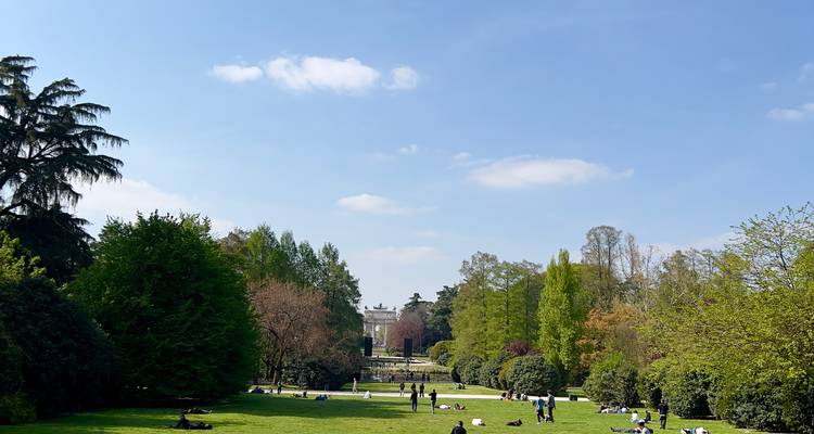 Vaste étendue herbeuse du Parco Sempione avec des personnes prenant un bain de soleil et l'Arco della Pace en arrière-plan.
