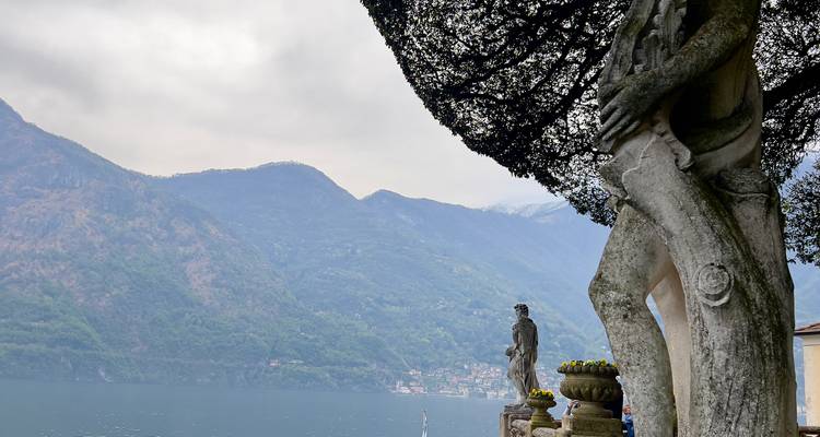 Des statues classiques et un grand arbre taillé surplombant le lac de Côme sous un ciel nuageux.