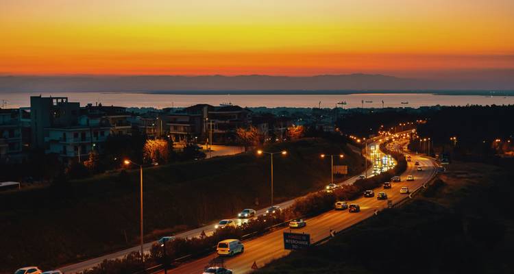 Le crépuscule doré illumine une autoroute animée qui s'incurve vers la baie de Thessalonique avec des montagnes lointaines en silhouette.