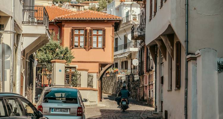 Un motocycliste roule dans une ruelle pavée étroite flanquée de maisons historiques aux teintes chaudes à Kavala.