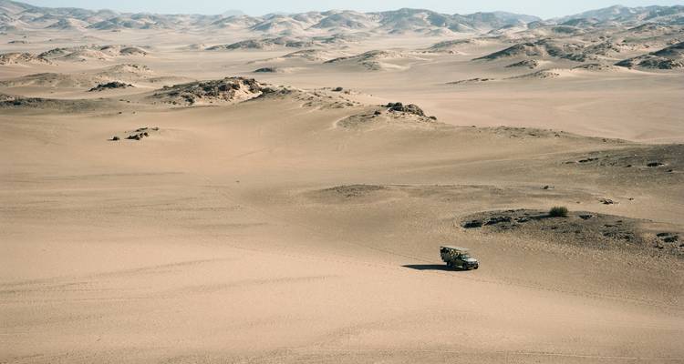 A lone overland truck traverses expansive rolling sand dunes under a hazy desert sky