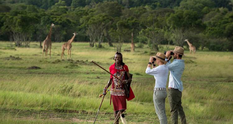 Two tourists with Maasai guide observing distant giraffes on an open grassy savanna.