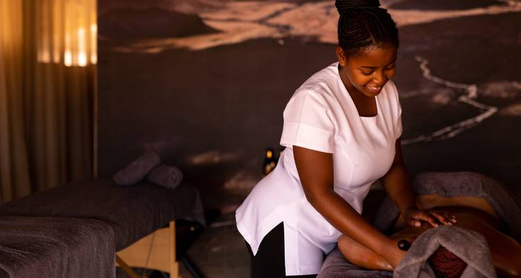 Smiling therapist giving a relaxing massage in a softly lit spa treatment room with dark linens.