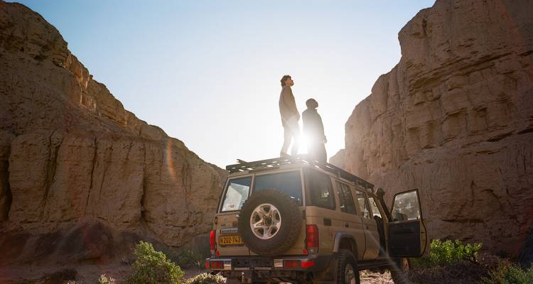 Two travellers standing on the roof rack of a safari jeep between high canyon walls, sun flaring behind them.